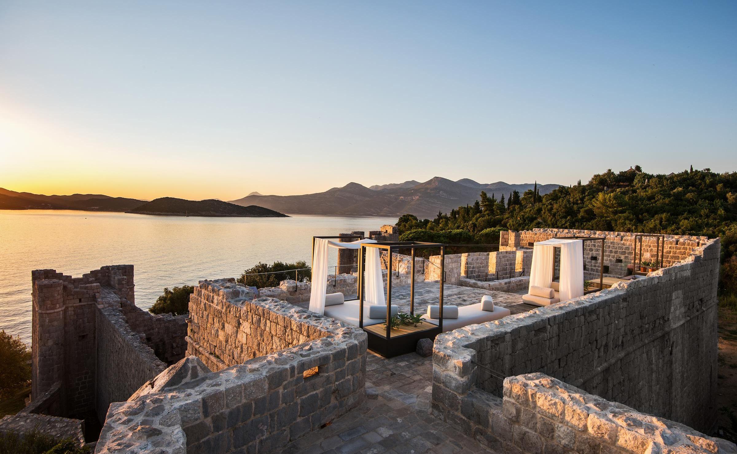 Contemporary daybeds with white curtains on a stone fortress roof with a panoramic view of the sea and mountains.
