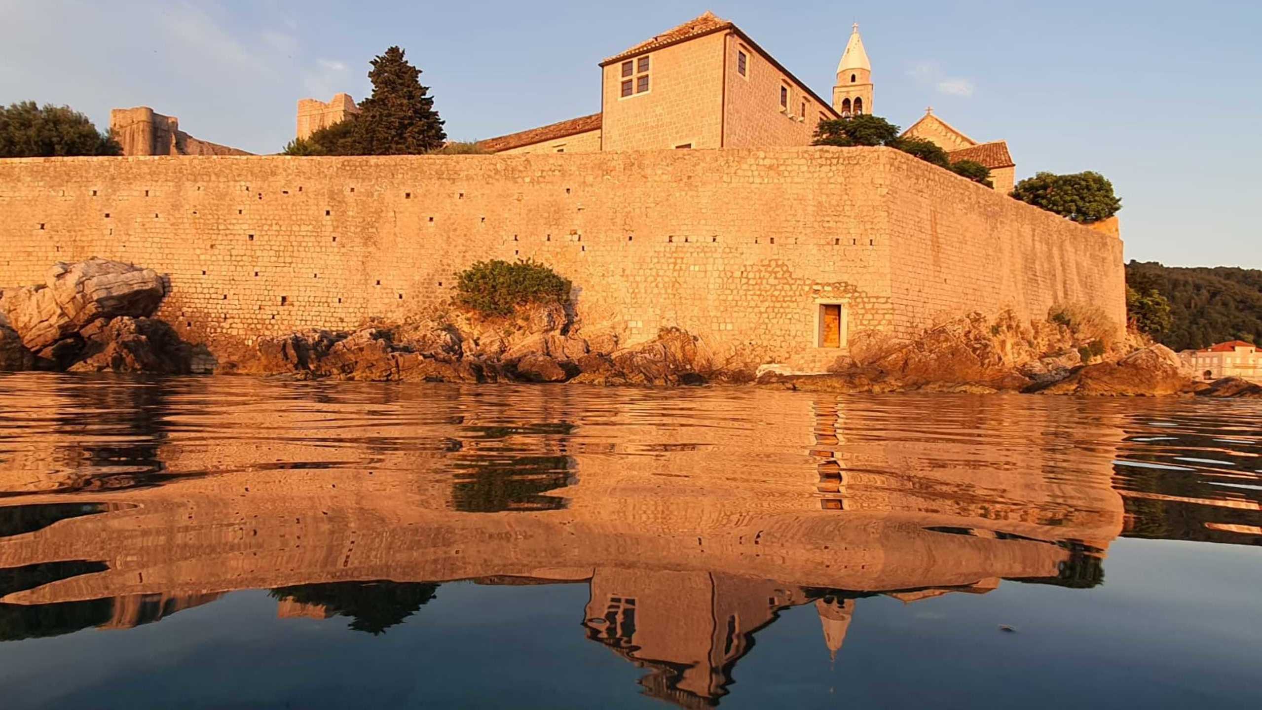 A tall stone wall with a small wooden door reflected in the calm surface of the sea during sunset.