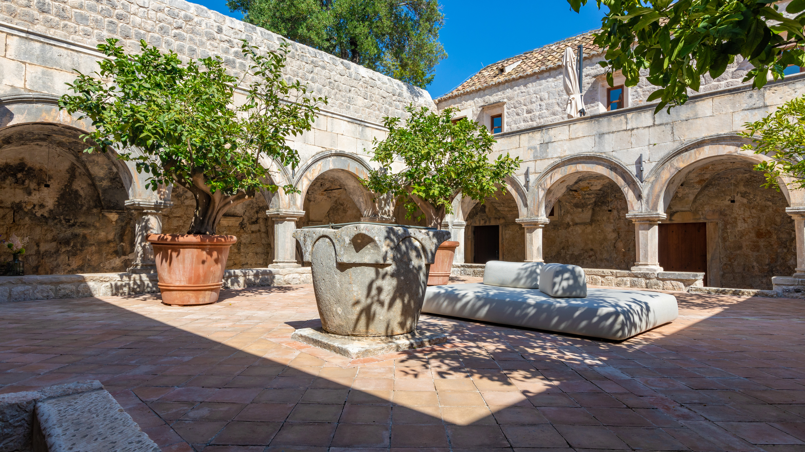 Sunlit courtyard with stone arches, a central well, and a large white outdoor sofa on a tiled floor.