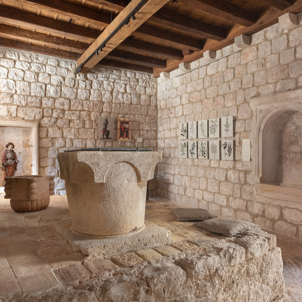 A rustic room with stone walls, wooden ceiling beams, a large stone basin, and framed botanical prints.