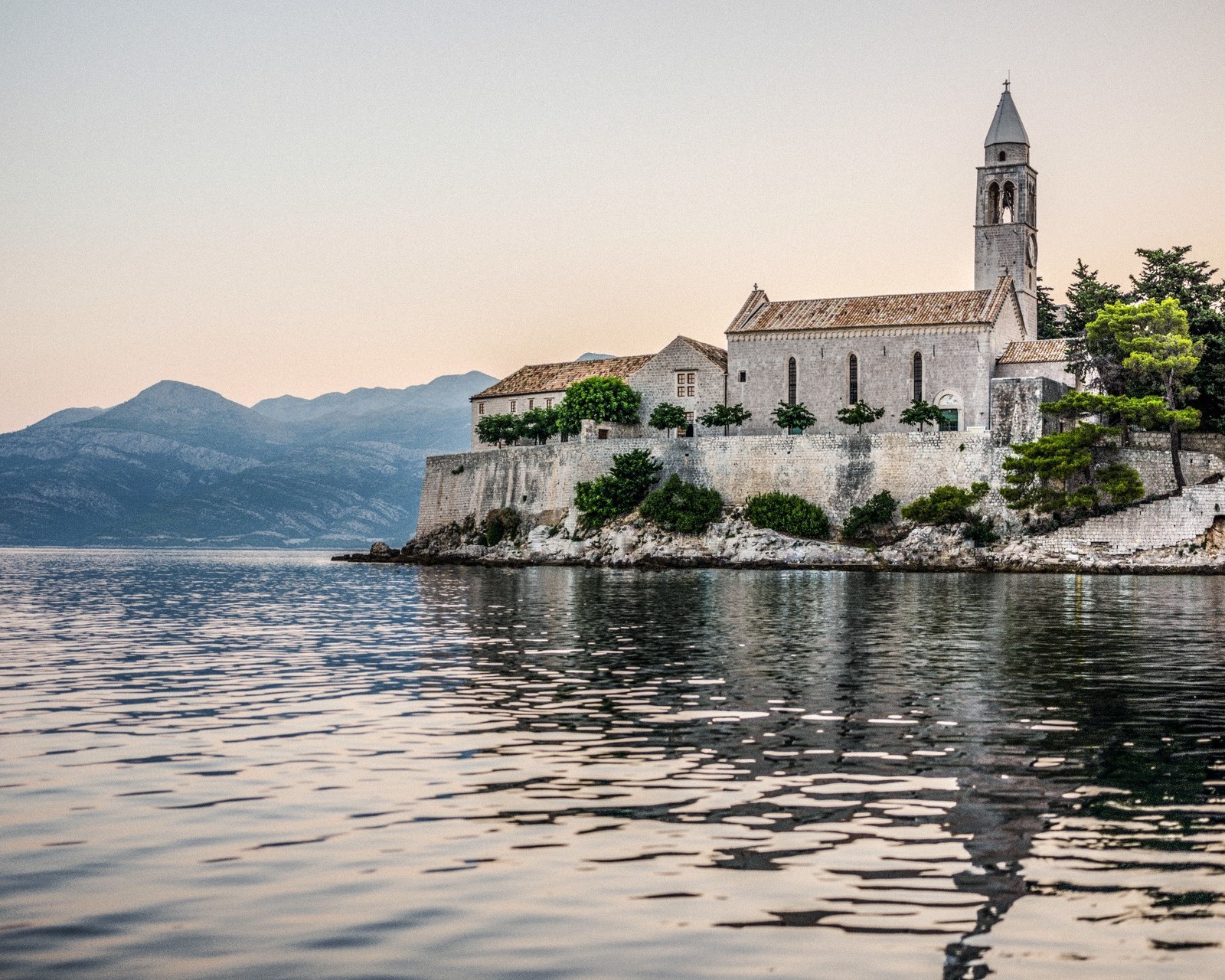 Stone monastery with a tall bell tower sits on a rocky cliff above the calm Adriatic sea at dusk.