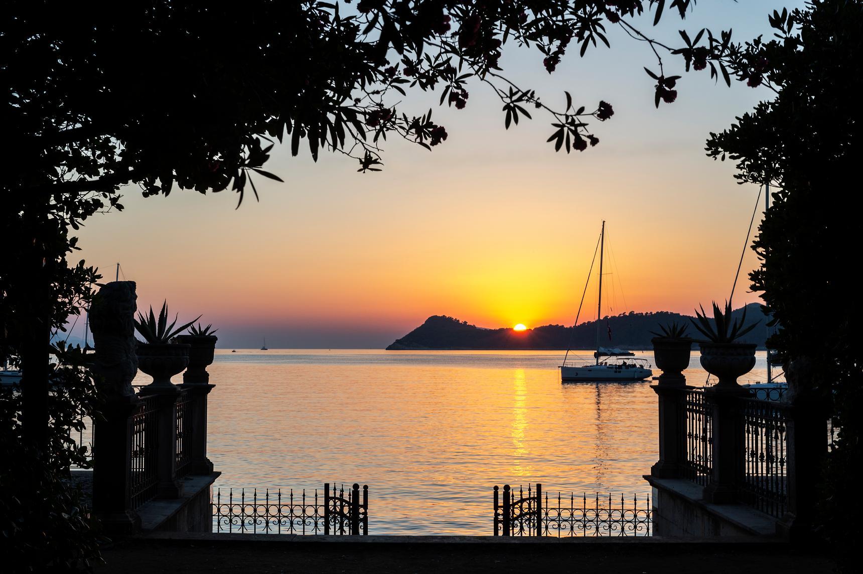 The sun sets over a calm sea and distant islands, framed by dark trees and a decorative stone gateway.