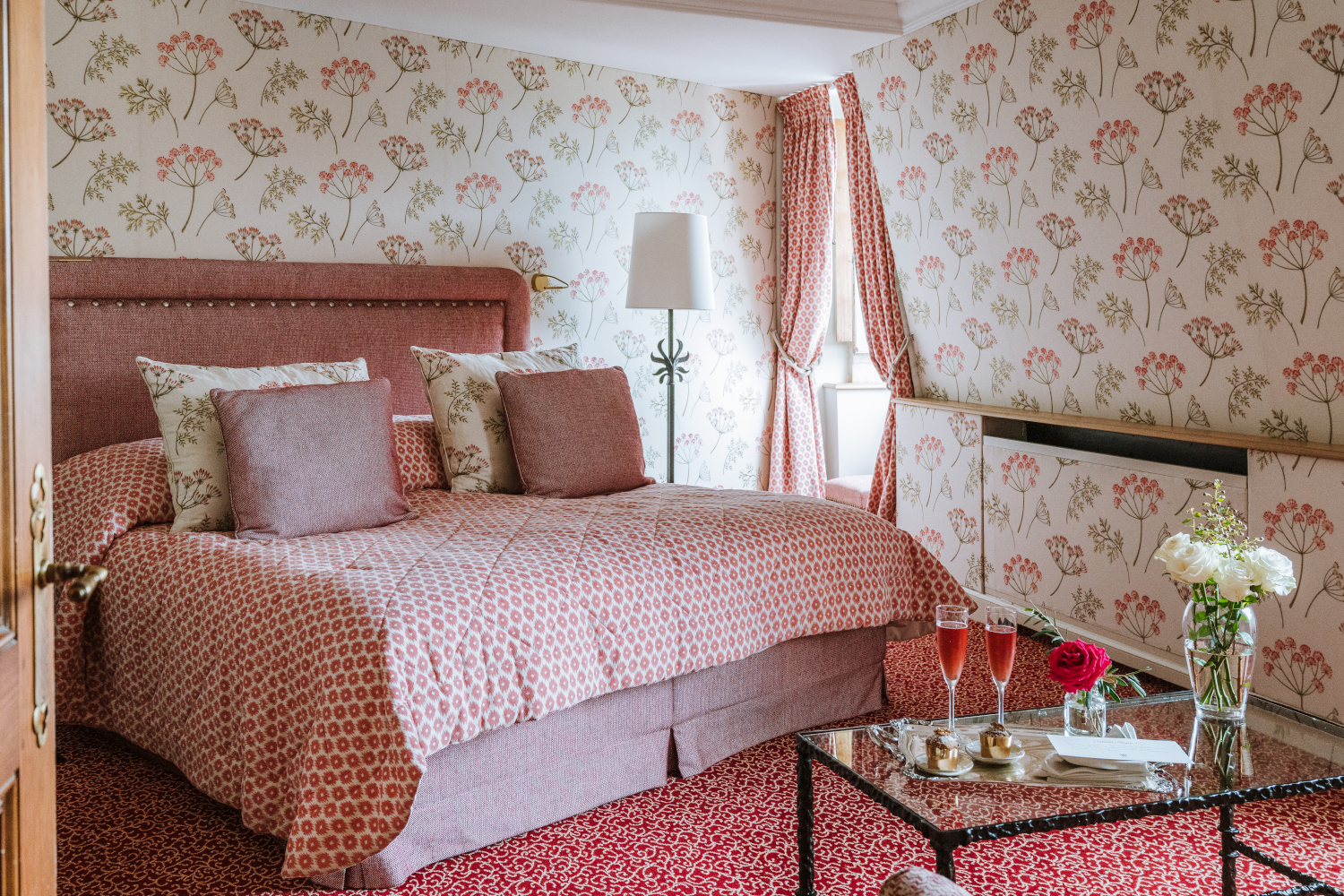 Bedroom interior with a bed with light red bedding, red carpeting and period wallpaper, and a small glass table in the foreground with drinks and flowers.