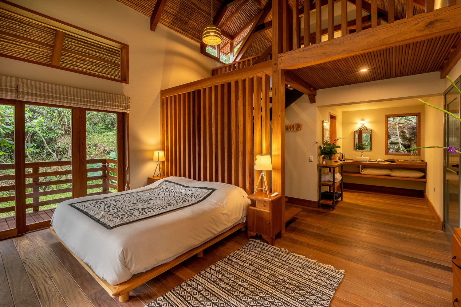 Bedroom interior with a double bed and natural textiles, wood floors and panelling, warm lighting and a view out to the green forest.
