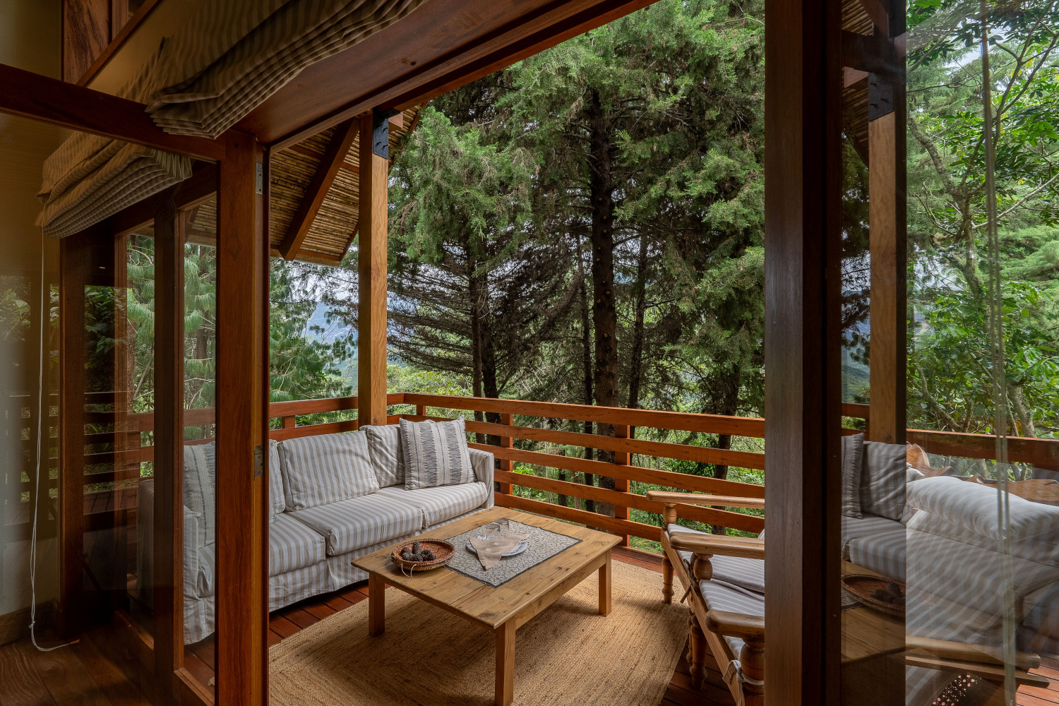 View through a door to an exterior terrace with light wood furnishings and linen upholstery and tall green trees just beyond the balcony.