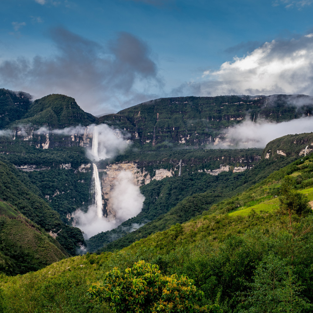 Long-distance view of Gocta Falls cascading down a green mountainside with clouds dotted around it.