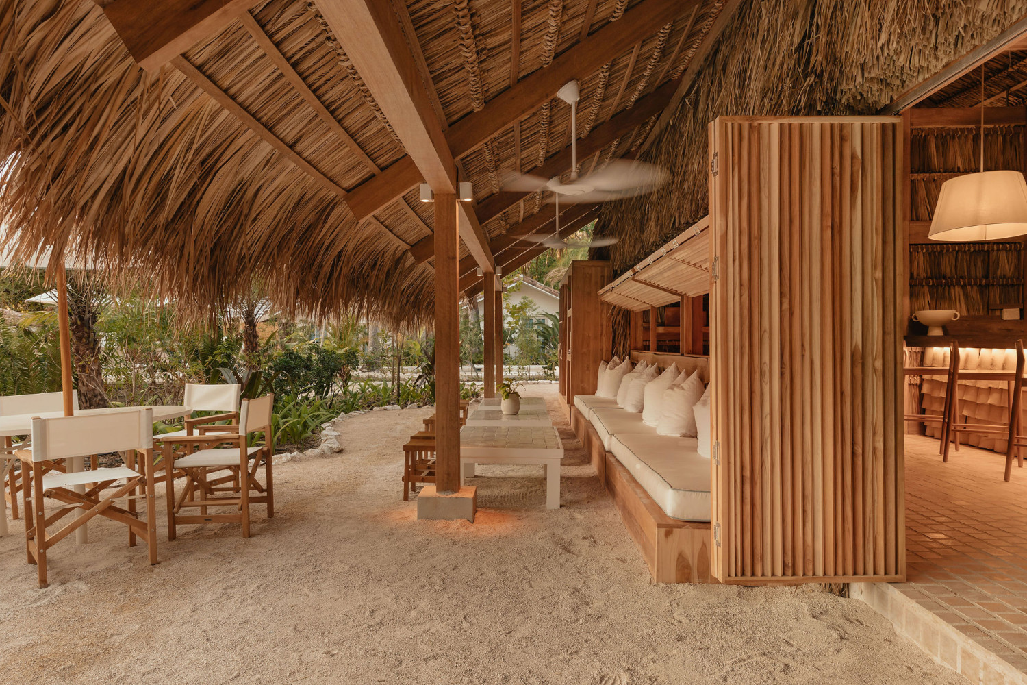 Lounge area with covered thatch roof and ceiling fans and sand floors, with banquettes, tables and a bar.