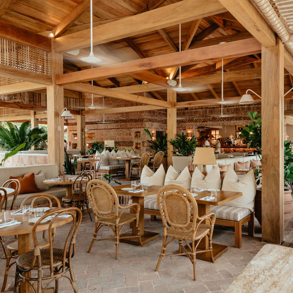 Interior of the restaurant with wicker chairs and tables, light wood beams, scattered green plants and stone floors.