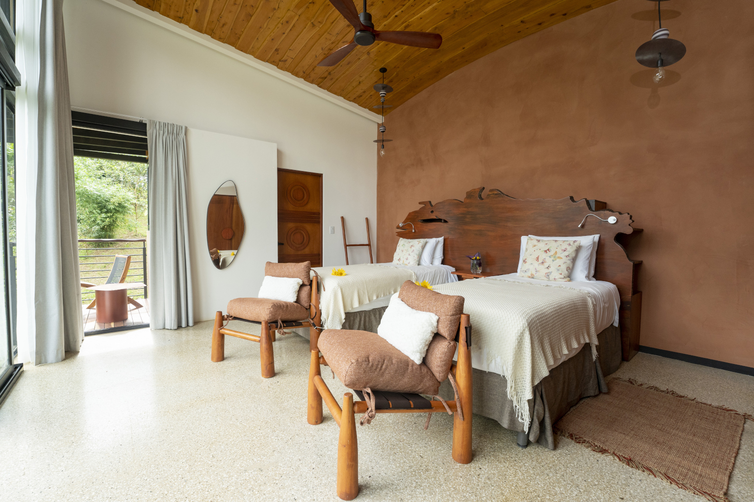 Interior of a bedroom with two beds with white sheets, brown wood walls, two wooden chairs chairs in front of the beds and a ceiling fan overhead.