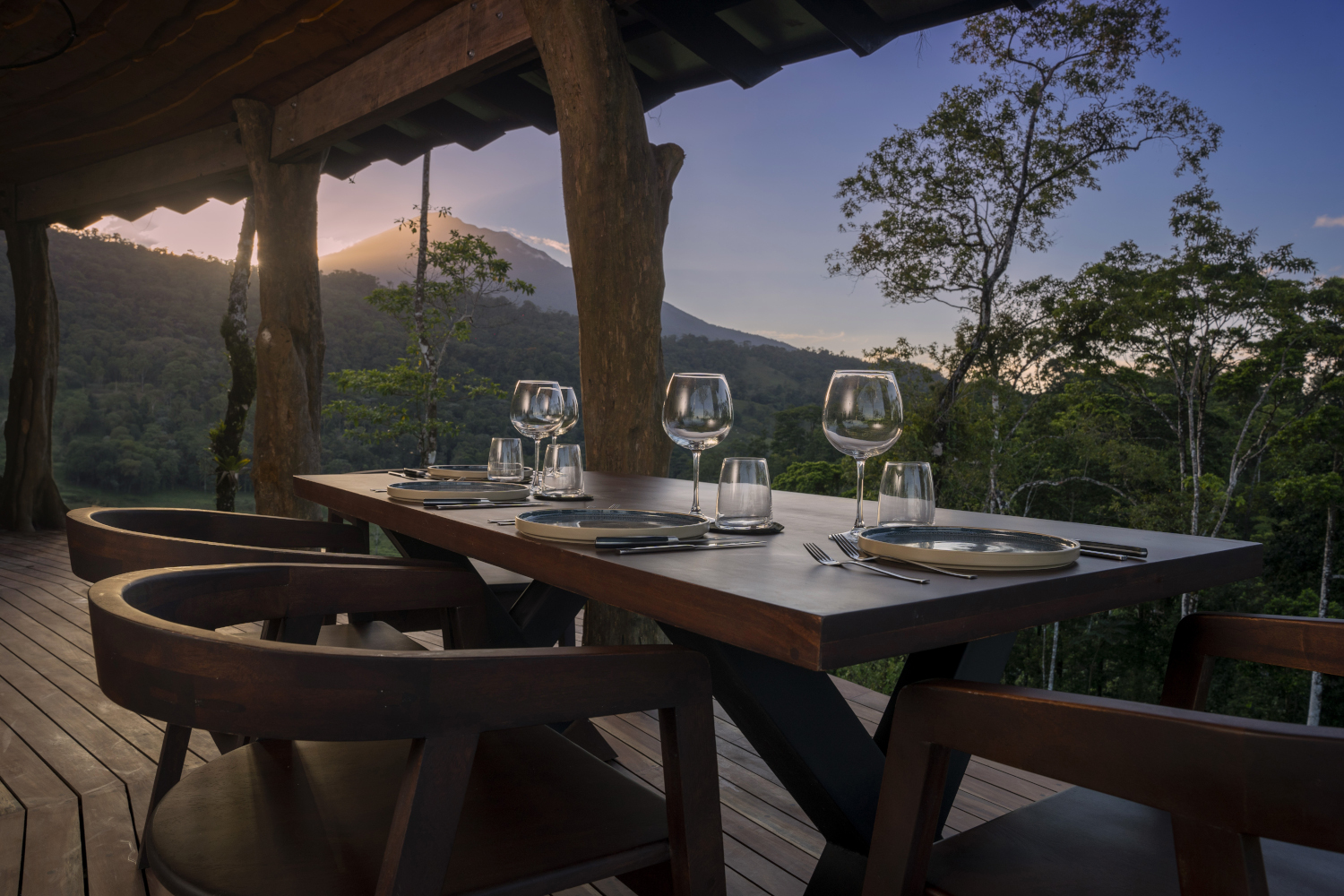 A wooden table on a covered terrace set with empty wine glasses, and in the background, trees, a pink sunset and volcano peak.