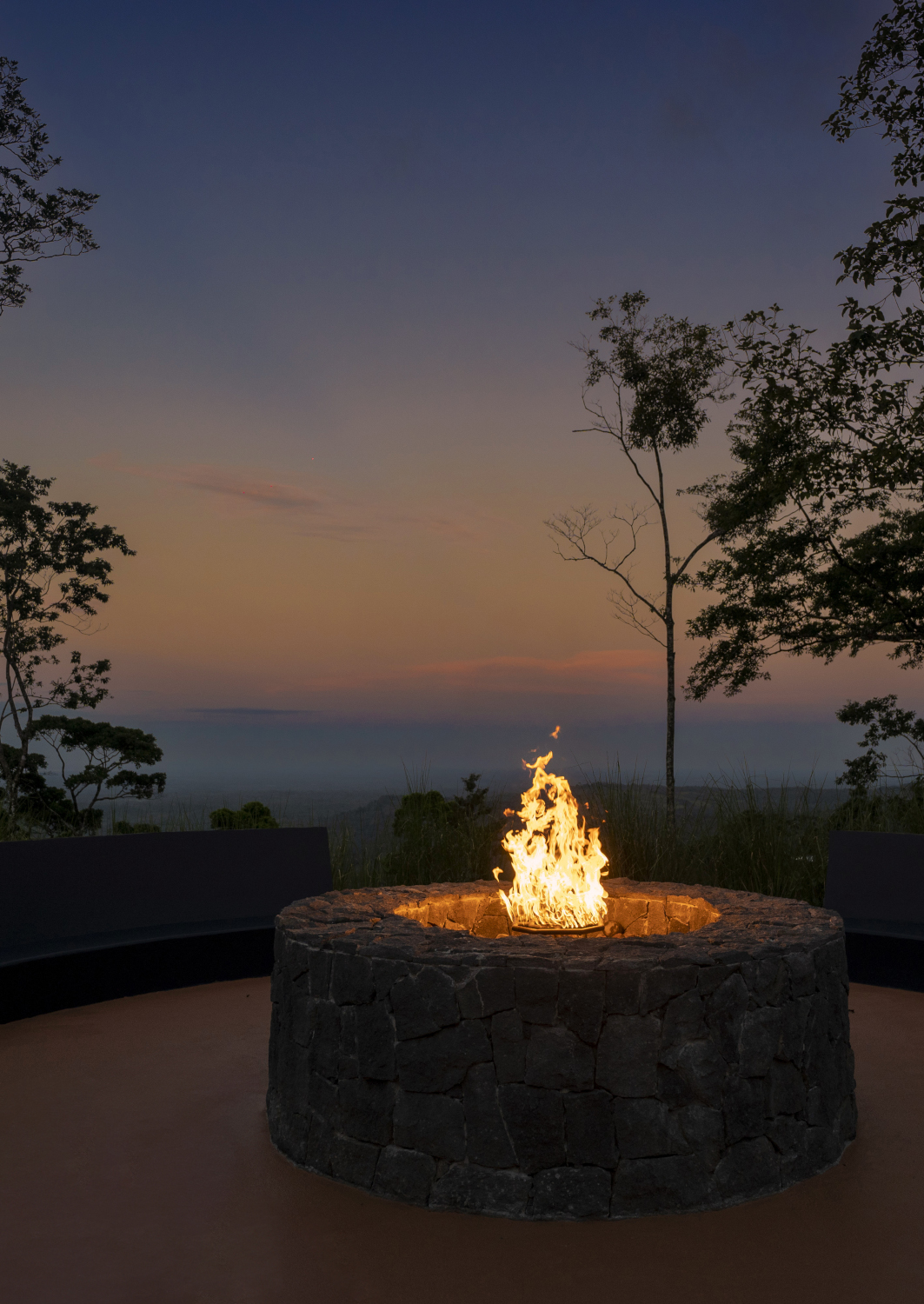 Lit firepit on a high terrace overlooking a darkening sunset sky with a few trees in the foreground.