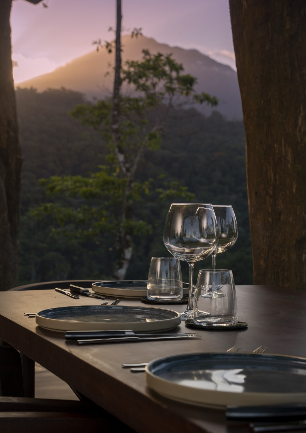 A wooden table on a covered terrace set with empty wine glasses, and in the background, trees, a pink sunset and volcano peak.