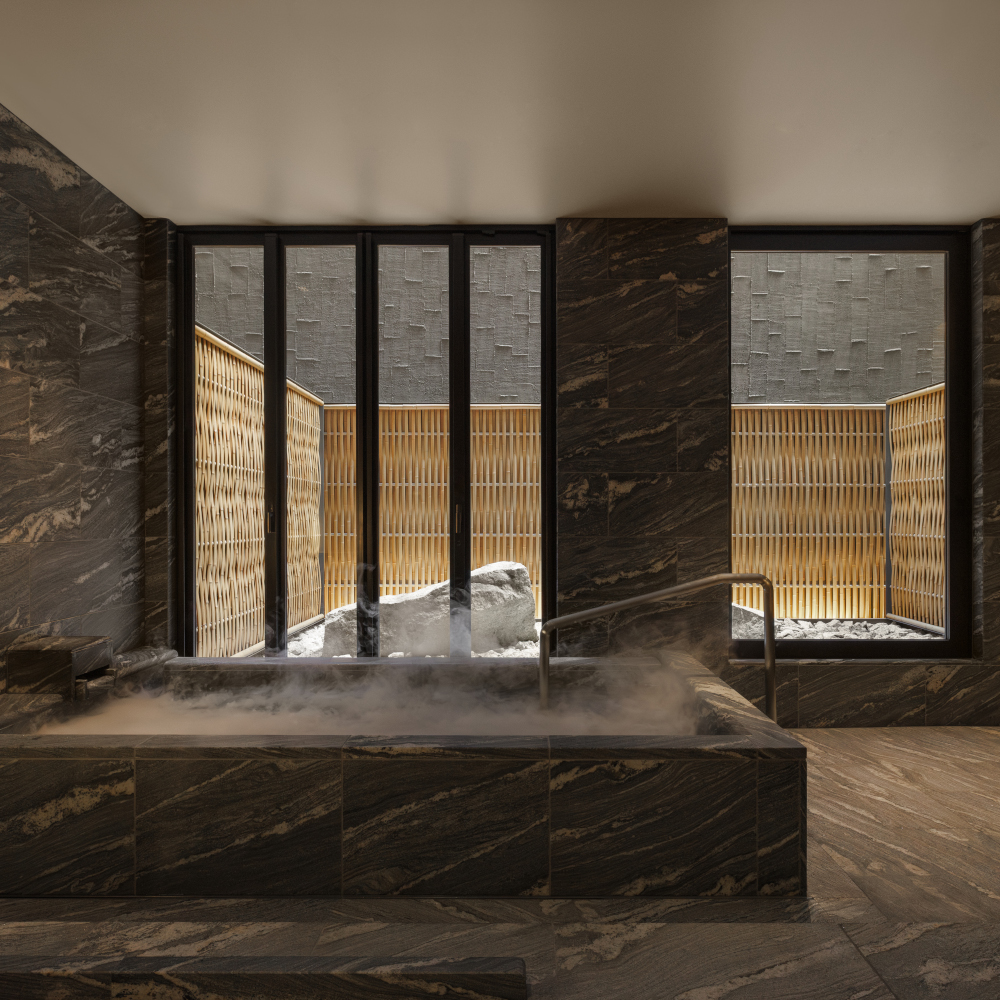 Private onsen tub steaming with water; the bathroom has dark tiles and there are windows overlooking an outdoor bathing terrace surrounded by a bamboo or rattan fence.
