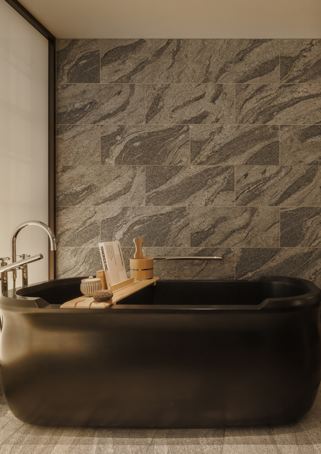 Dark stone soaking bathtub with a tray of toiletries in a room with grey stone tiled walls.