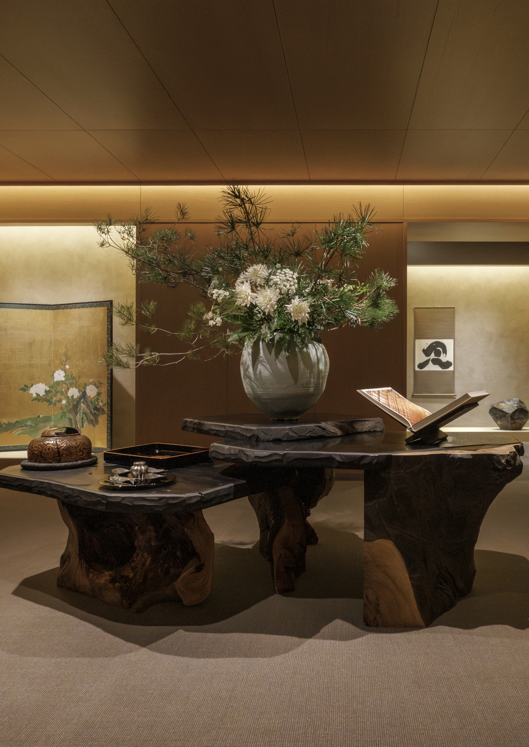 Lobby reception with a welcome table made of wood fossils on top of which are a large vase with a floral arrangement and a decorative book open on a stand.