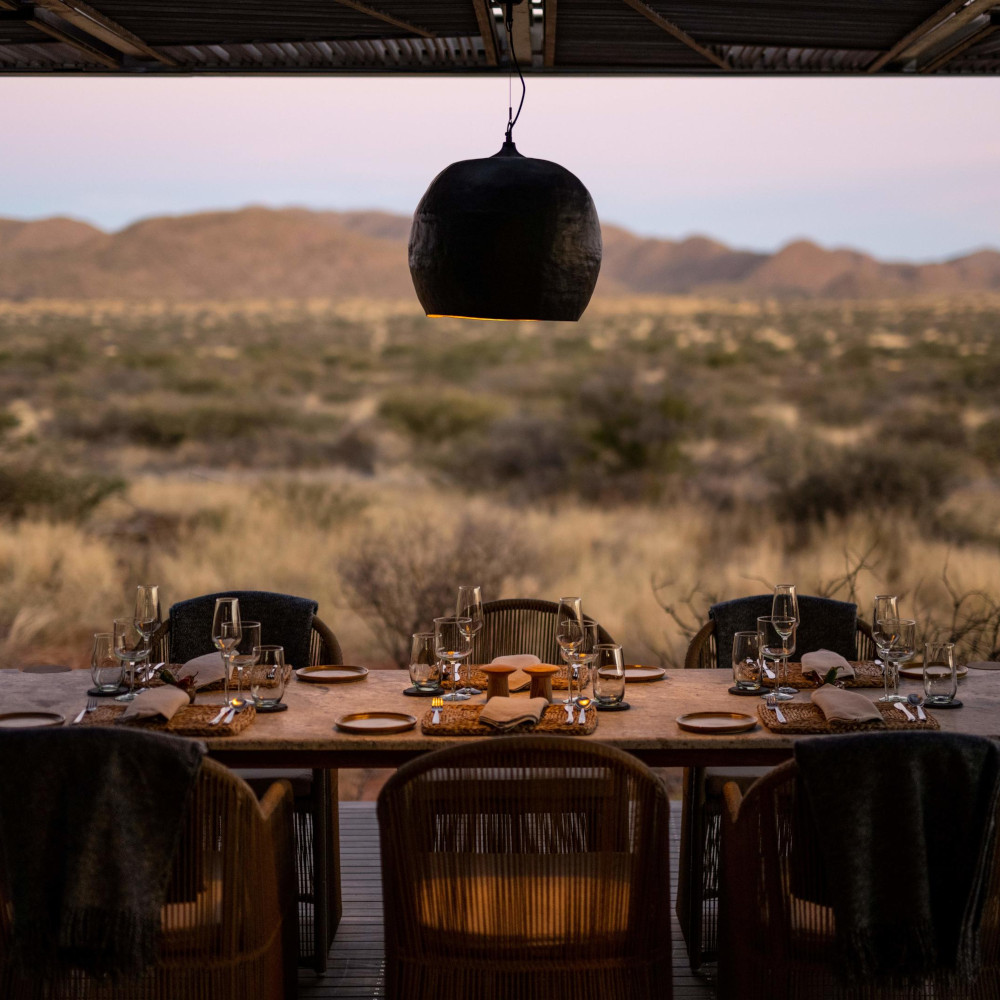 Six chairs at a table set with dinner plates and wine glasses with a round black hanging lamp shade above and beyond an open-air view of the bush and mountains.