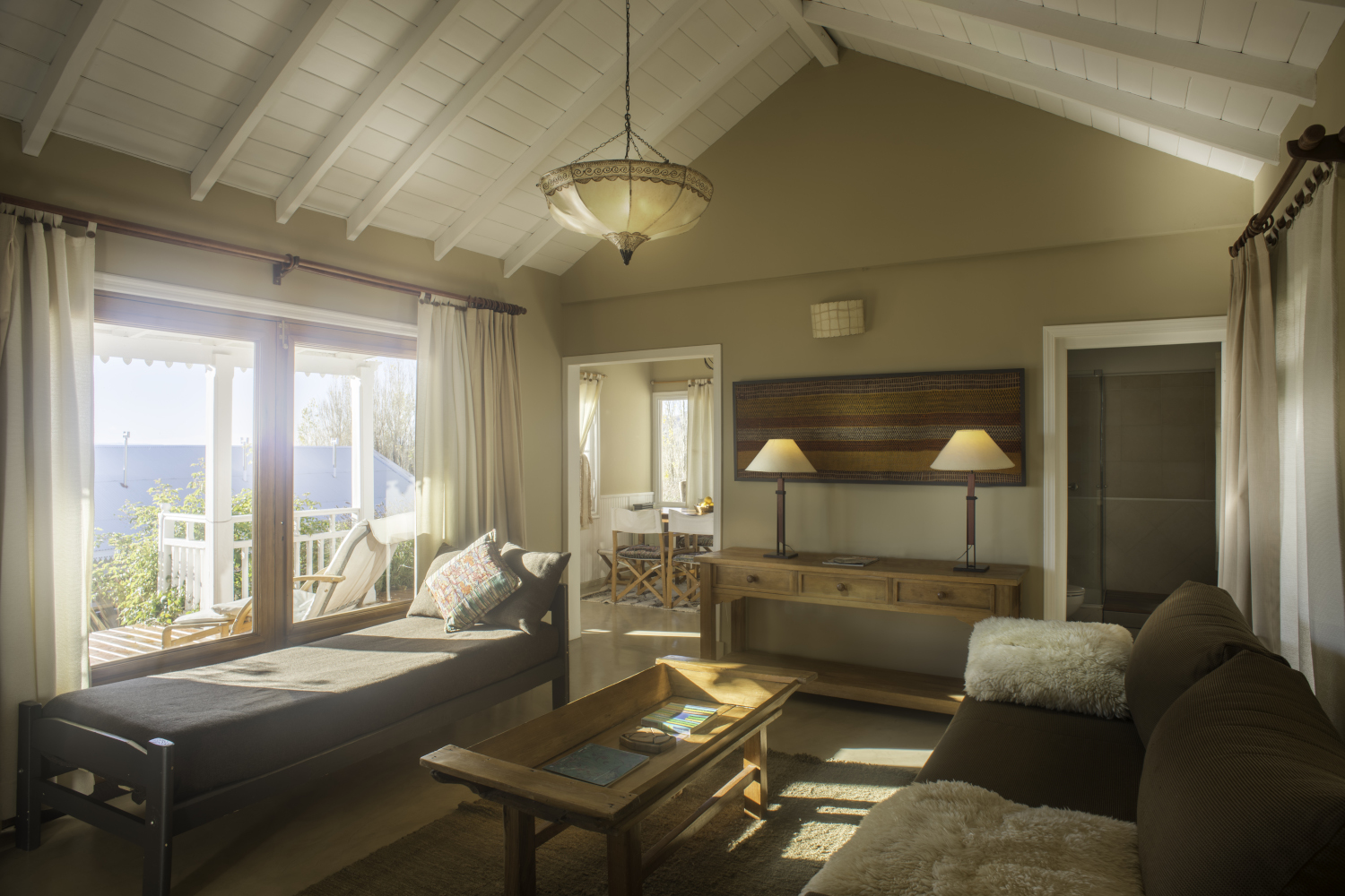 Living room area with big windows overlooking the lake. Inside is a brown sofa with white throw pillows, a side desk with two lamps, a chaise lounge underneath the windows and a coffee table in the centre.