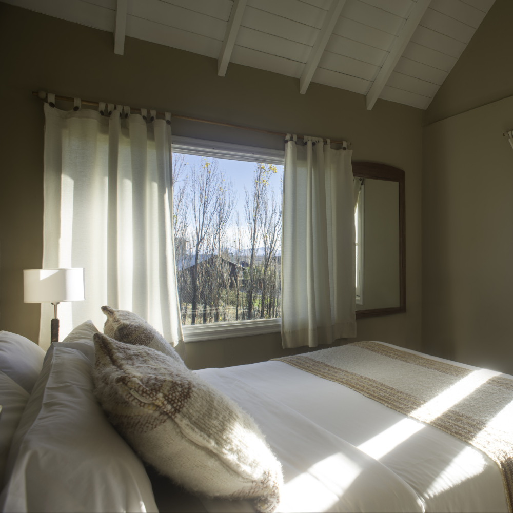 Double room dressed in white and beige bedding with a window and white curtains, and white timber ceiling.