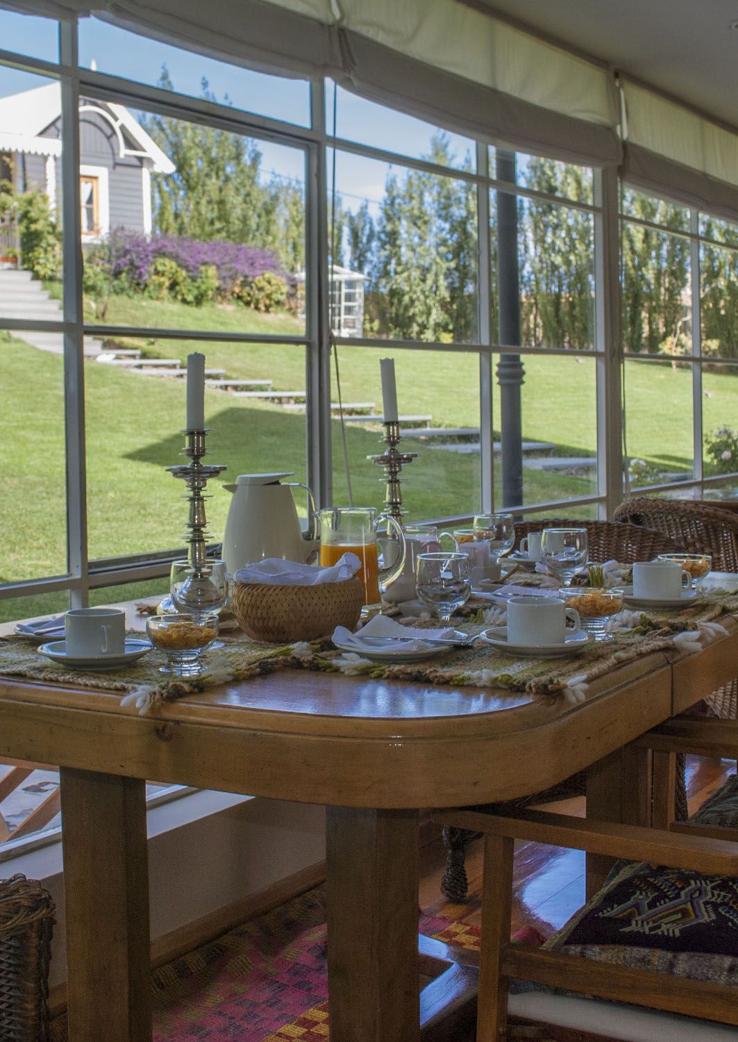 Wooden table set for breakfast with juices, coffees and plates, beside big windows looking out onto a green lawn.