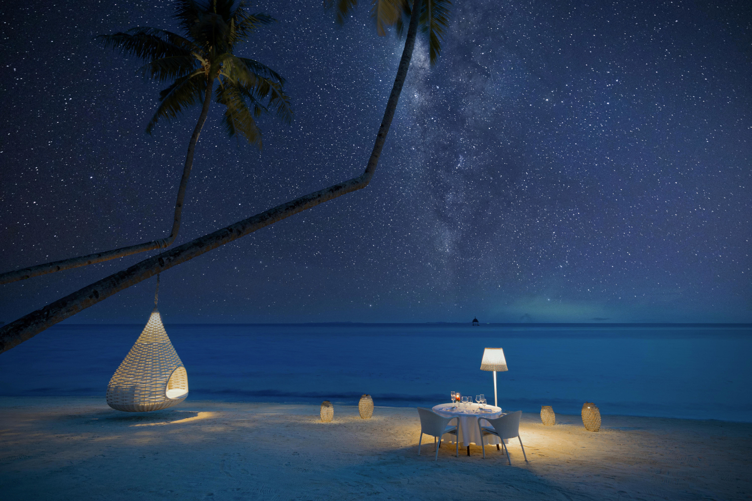 Starry sky with Milky Way over a beach table lit by a soft lamp next to the dark sea.