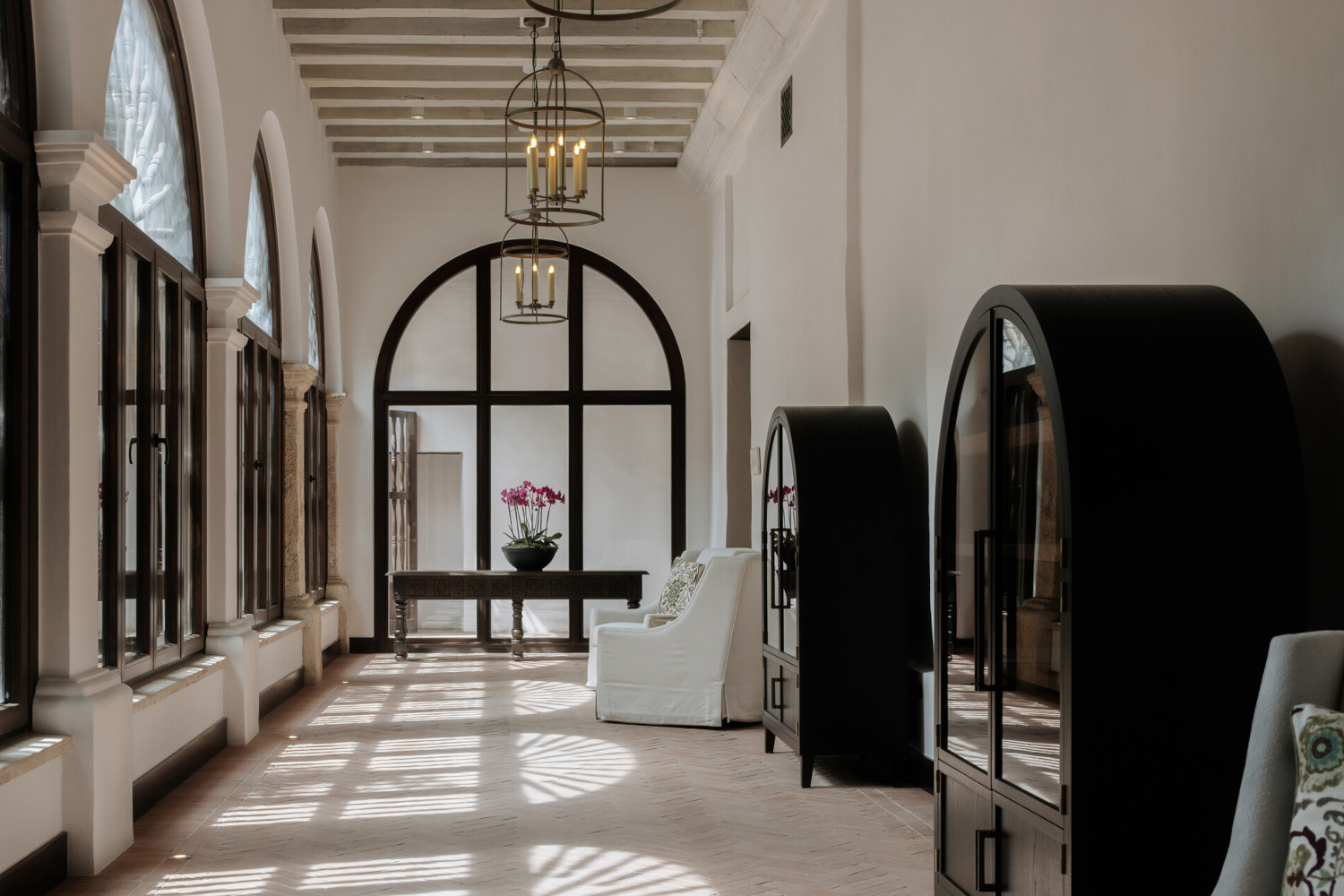 Interior corridor with high ceilings, white walls, antique furnishings and rounded historic windows.
