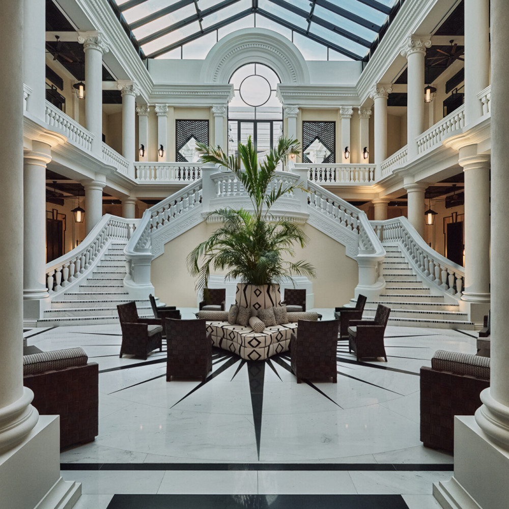Grand interior entranceway with white marble floors and a palm tree amid dark brown chairs, and two marble staircases leading up to a mezzanine area with skylight ceiling.