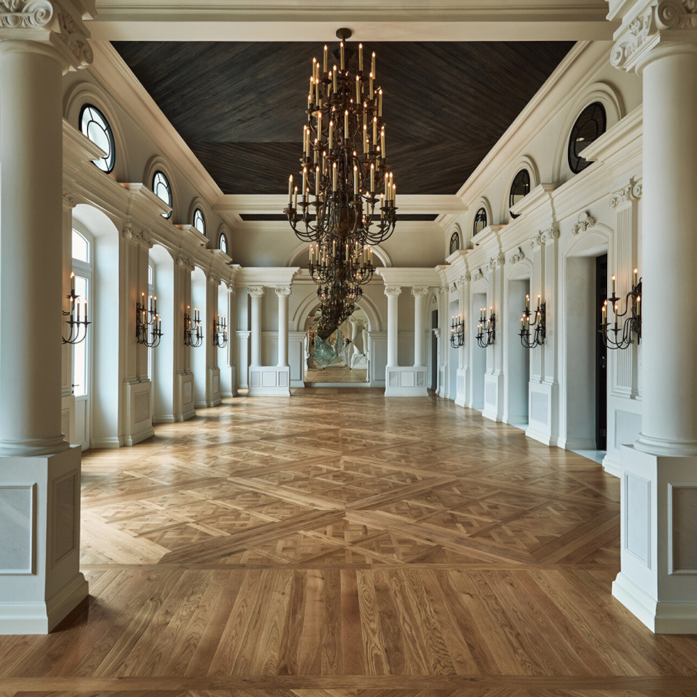 Empty ballroom with white marble columns and parquet wood floors and a long chandelier.