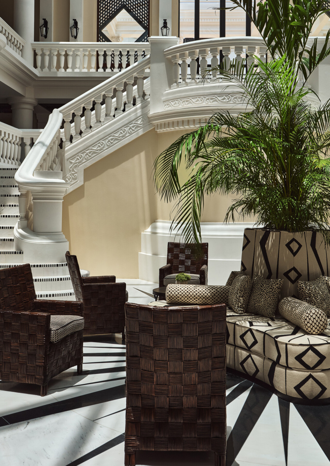 Grand interior entranceway with white marble floors and a palm tree amid dark brown chairs, and two marble staircases leading up to a mezzanine area with skylight ceiling.