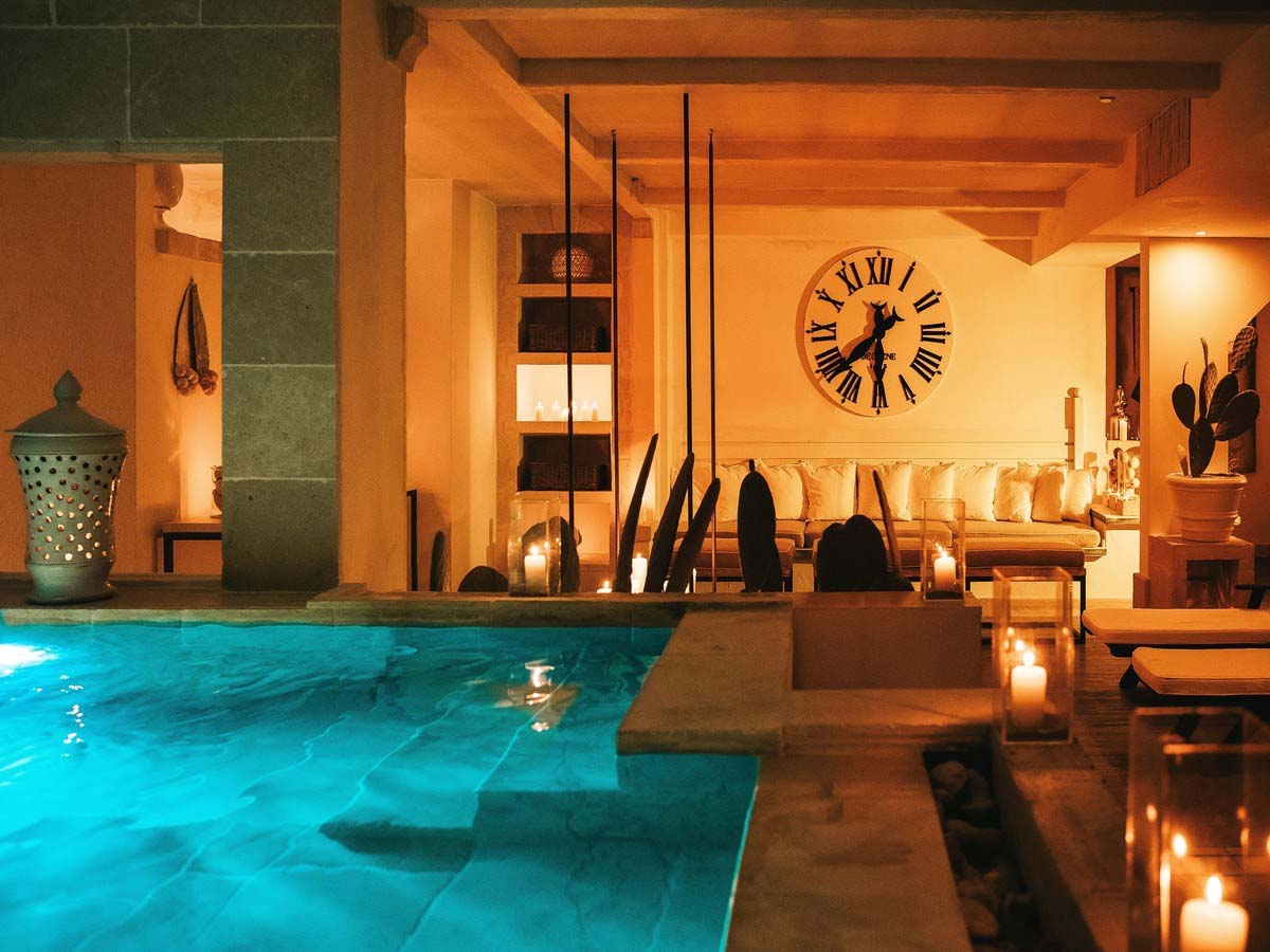 A spa setting with a thalassotherapy pool that has deep steps, in a candlelit space with lanterns, a long sofa with cushions and a big clock on the wall. The sauna is on the left behind the pool.