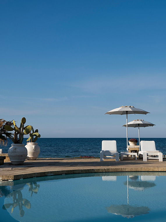 The curved edge of a swimming pool under blue skies with two sun beds, umbrellas, potted plants and sea views.