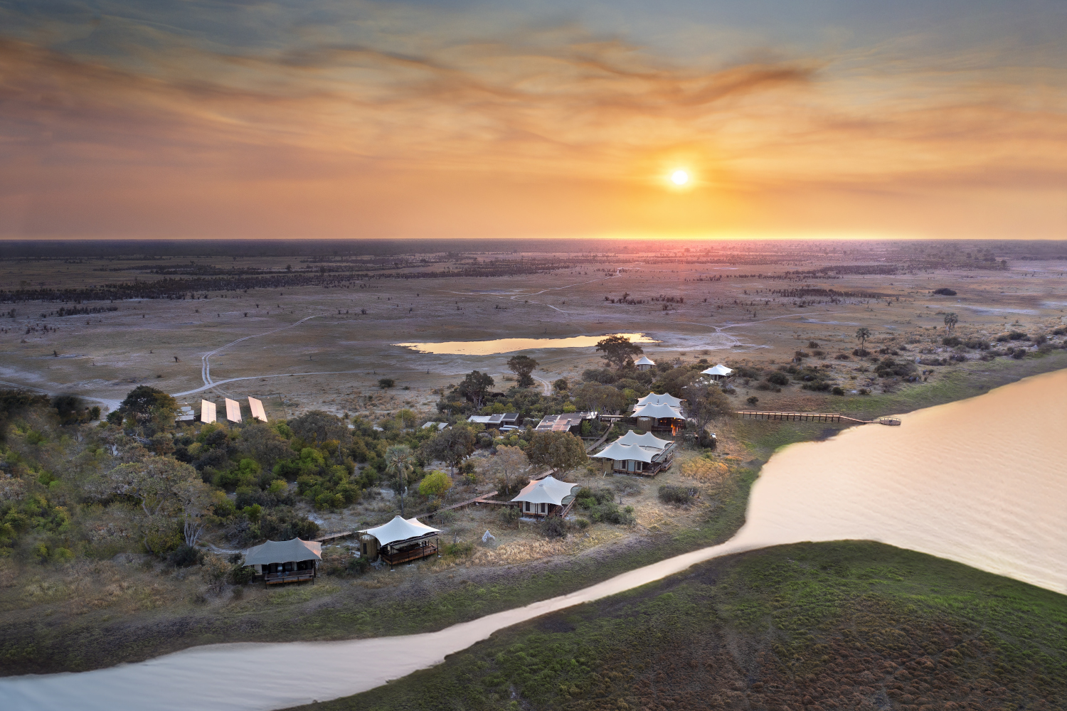 View of a tented safari camp from above with a lagoon flowing along the foreground and the bushland and a yellow sunset beyond.