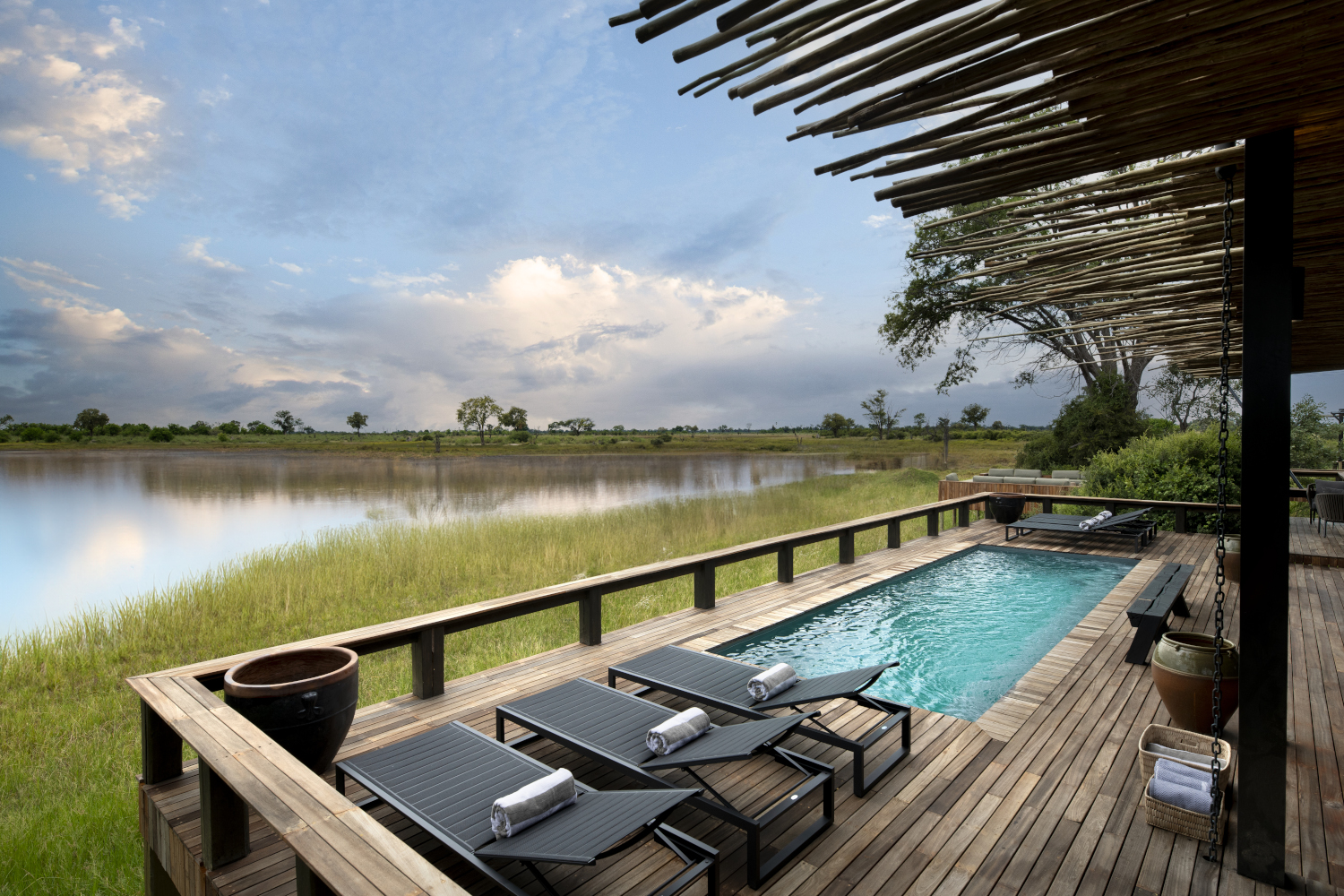 A blue swimming pool with three lounge chairs on a wooden deck and thatch overhand looking out over a wetland with water and grass.