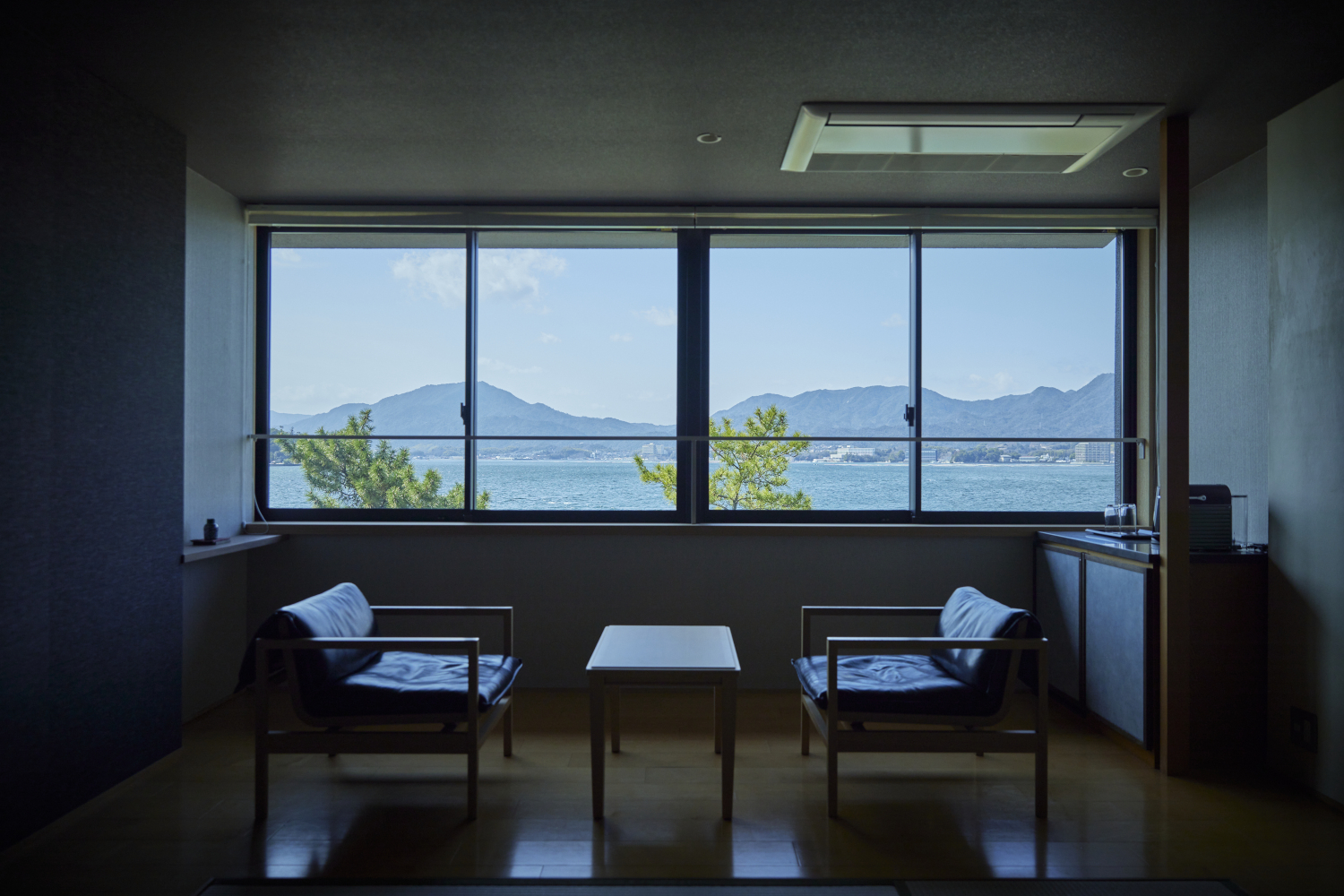 Interior view of a seating area with two vintage chairs facing a small coffee table in front of a long picture window overlooking the sea and mountains.