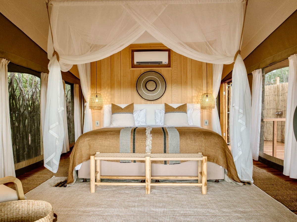 Interior of a safari tent showing a bed centered under a draped mosquito net with symmetric lighting and decor.