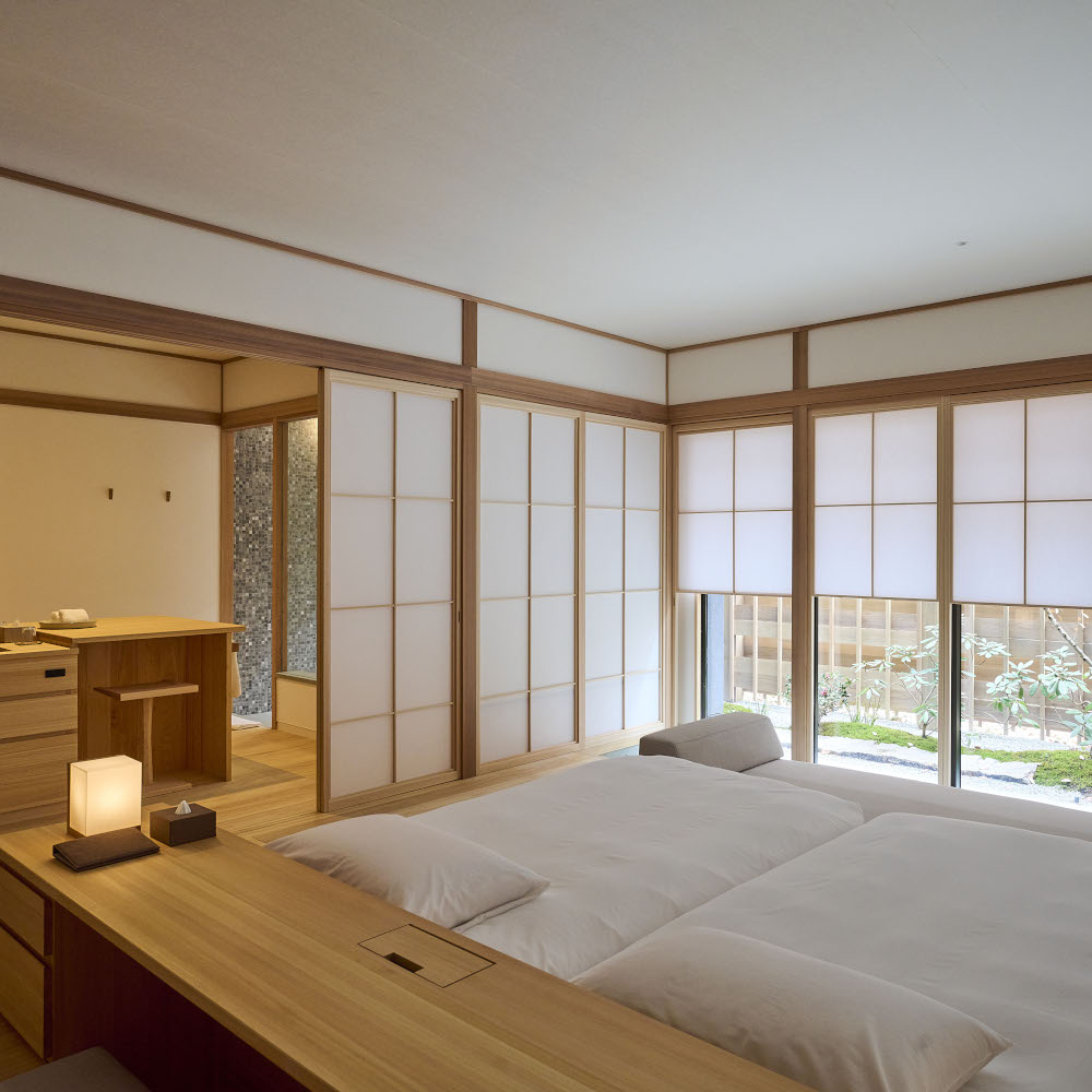 Interior of a bedroom with a minimalist bed with white bedding, light wood frame and white walls with light wood edging and accents, and large windows with paper screen panels.
