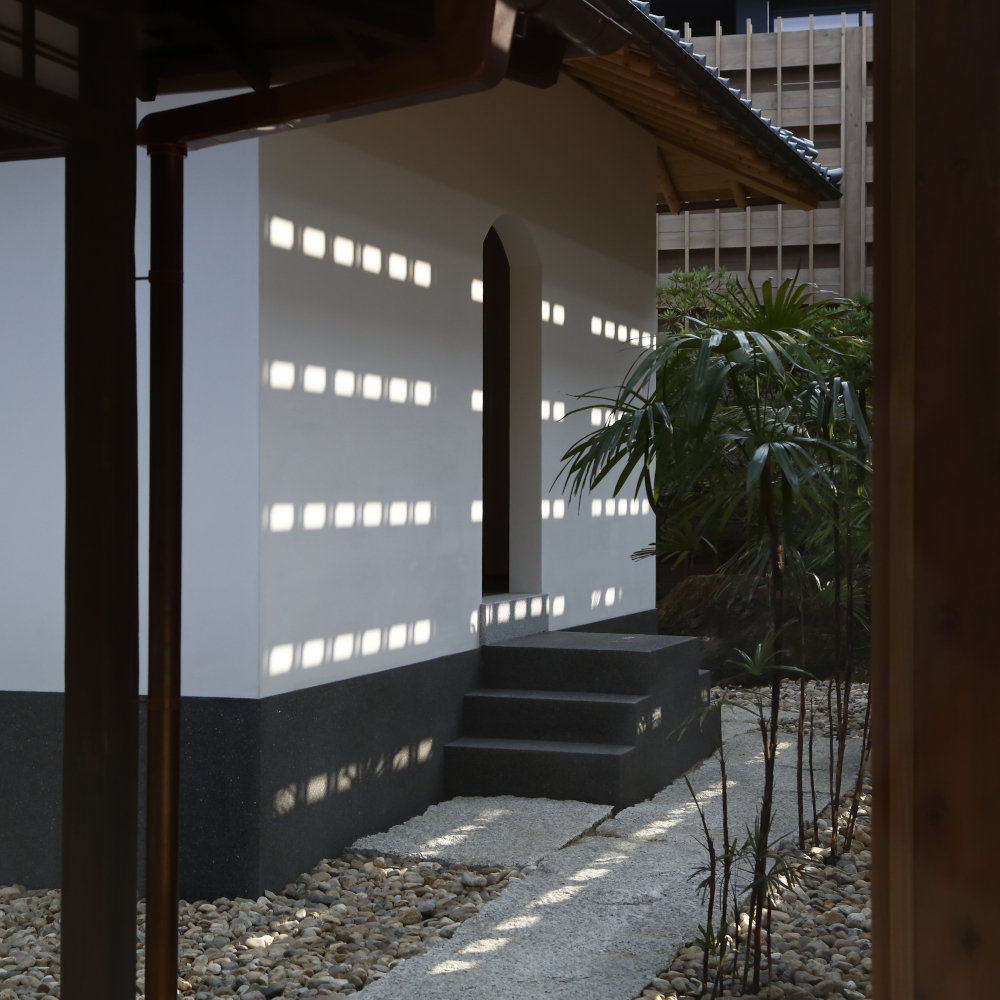 Exterior of an entryway with a rounded door and paved path surrounded by stones, entering into a white-washed building with tile roof.