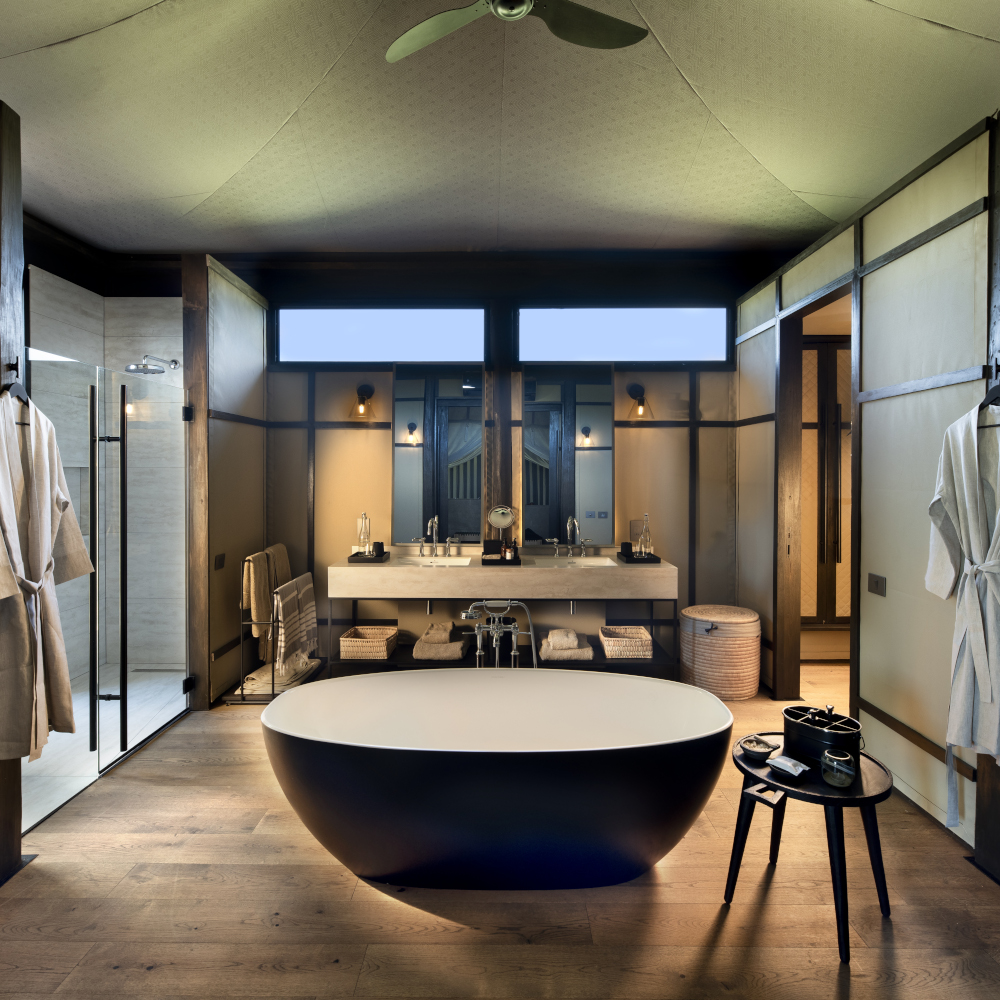 Interior of a safari tent bathroom with a freestanding black tub and small side table, and two bathrobes hanging up and sinks and mirrors in the background.