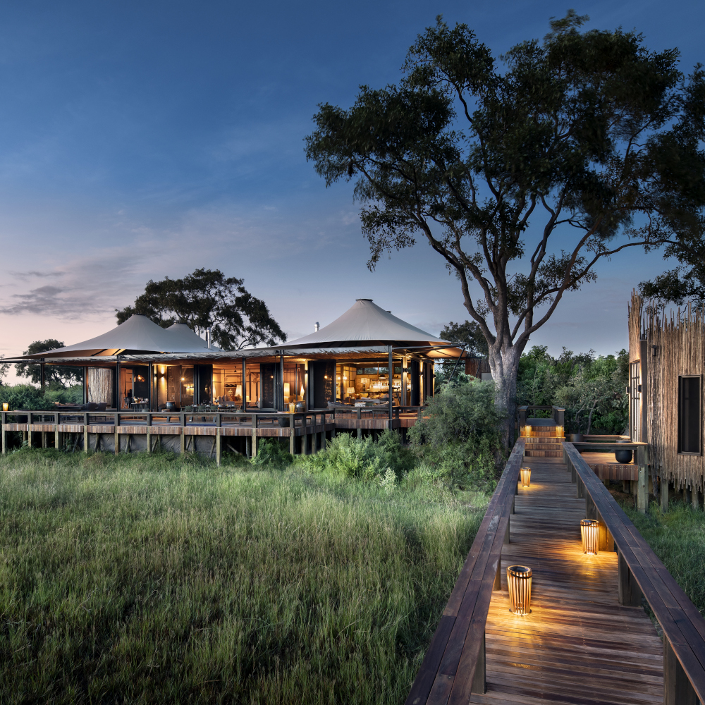 An evening sunset view of a tented safari camp with wooden decking and a wooden walkway lit by small amber lanterns.