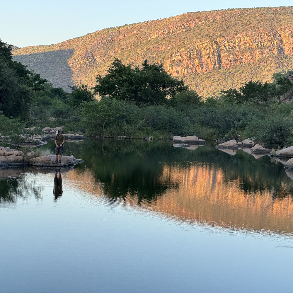 Man stands on a distant rock next to a large river or pool with trees in the near background and an illuminated mountain further distant.