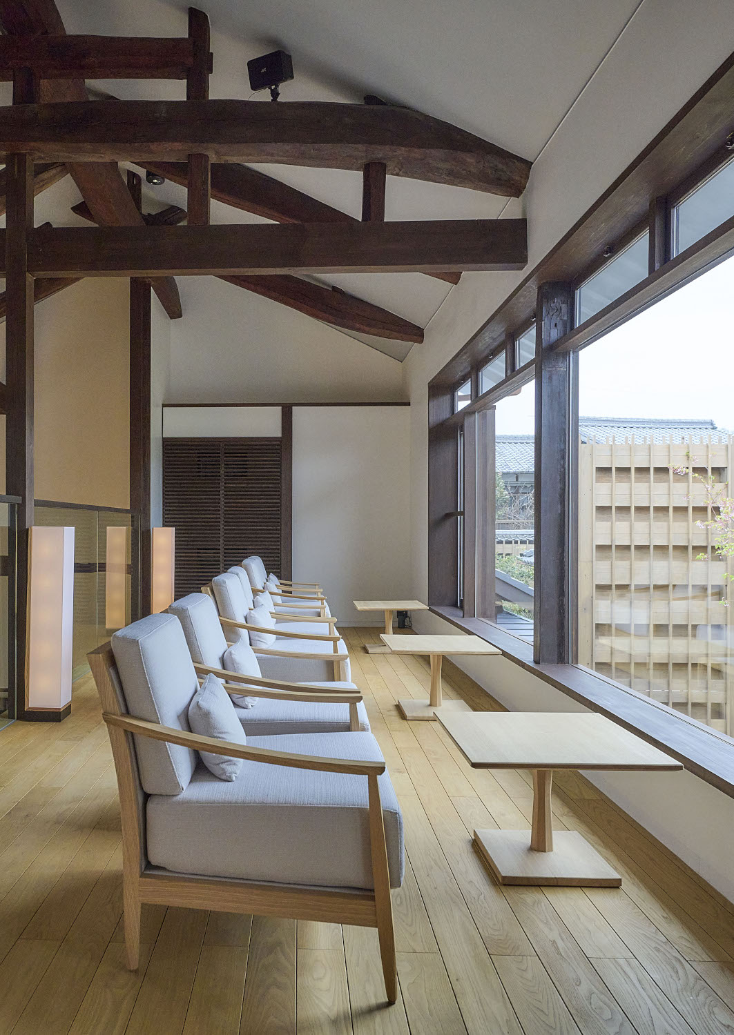 Interior of a lounge area with several white lounge chairs facing a few small wooden tables an a set of large picture windows looking out over buildings.