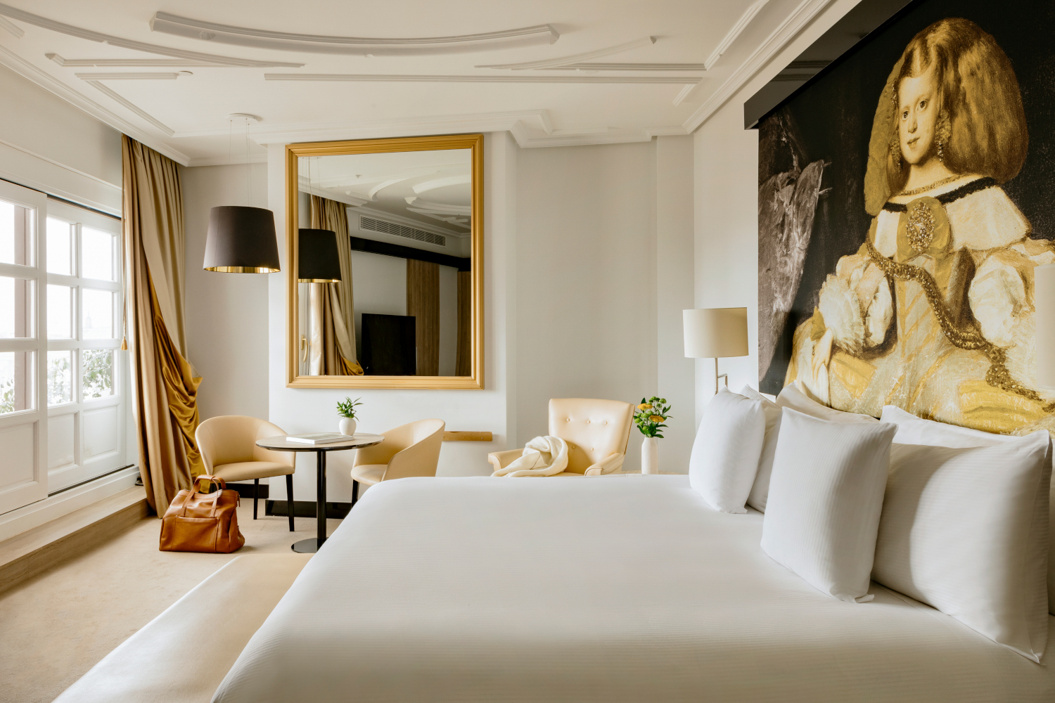Interior shot of a bedroom with a white bed in the foreground and a large mirror and lamp with a black shade against the back wall.