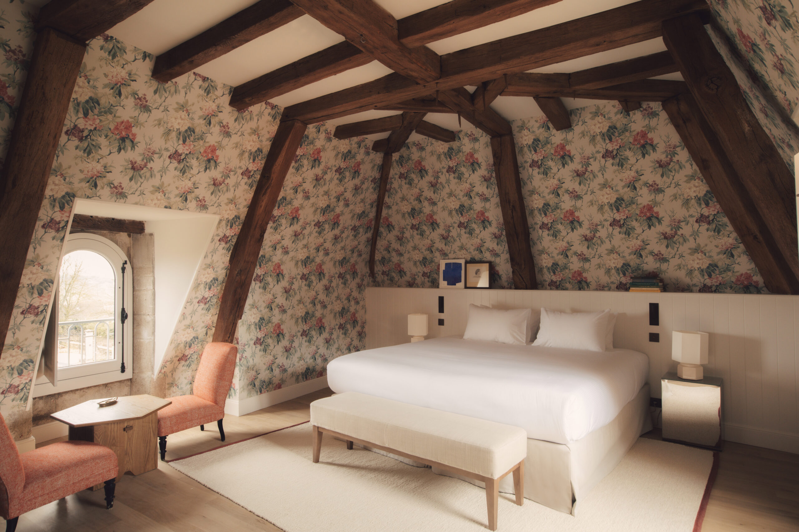 Bedroom with a bed with white bedding, floral wallpaper and dark wood beams.