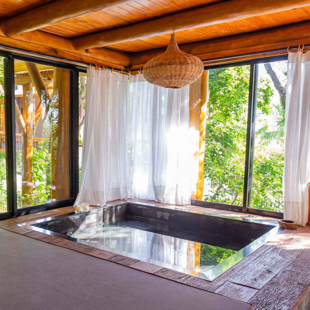 Interior of a small square plunge pool in a wooden room surrounded by floor-to-ceiling windows overlooking greenery.