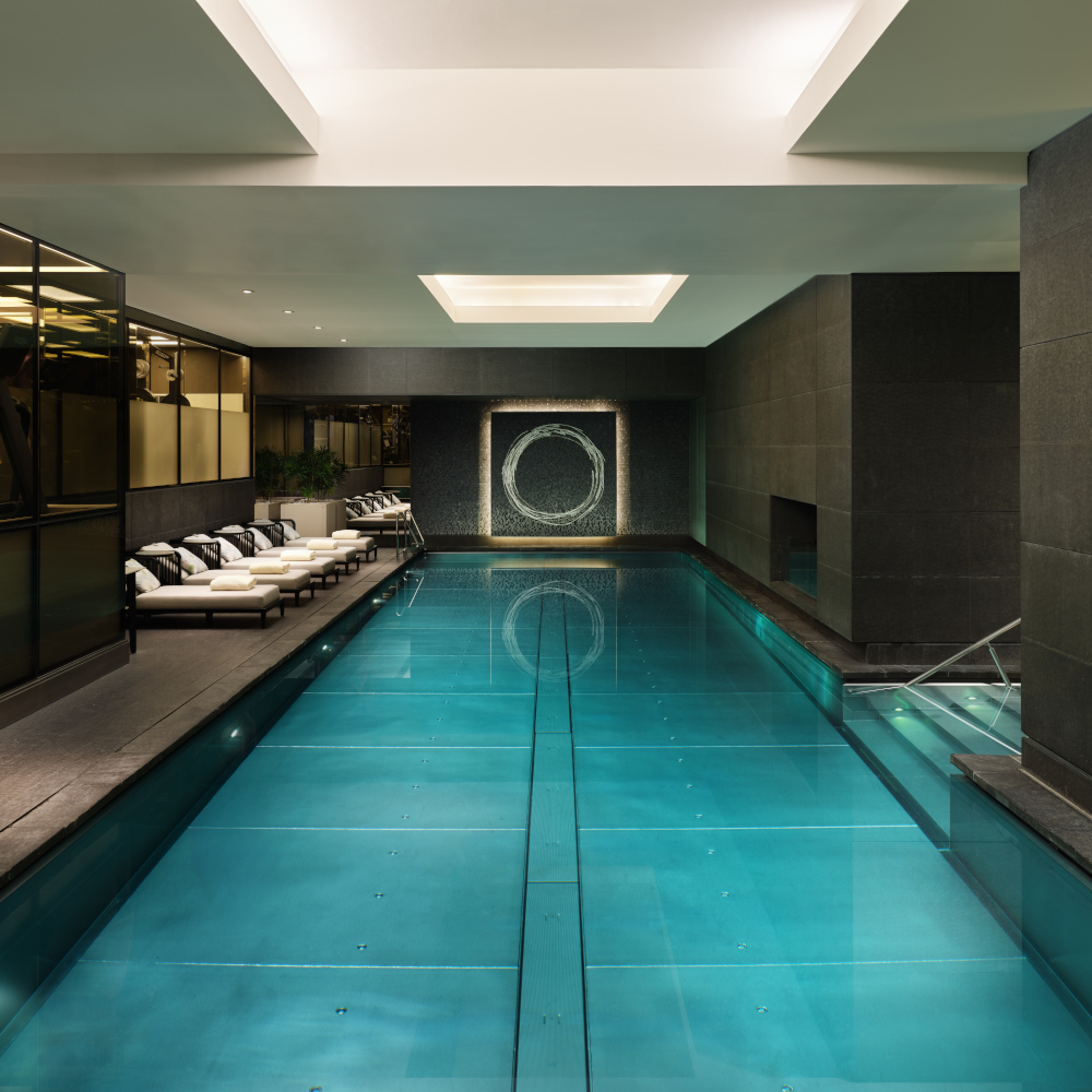 A long, rectangular indoor pool with dark grey walls and a line of white recliner chairs along the left side.