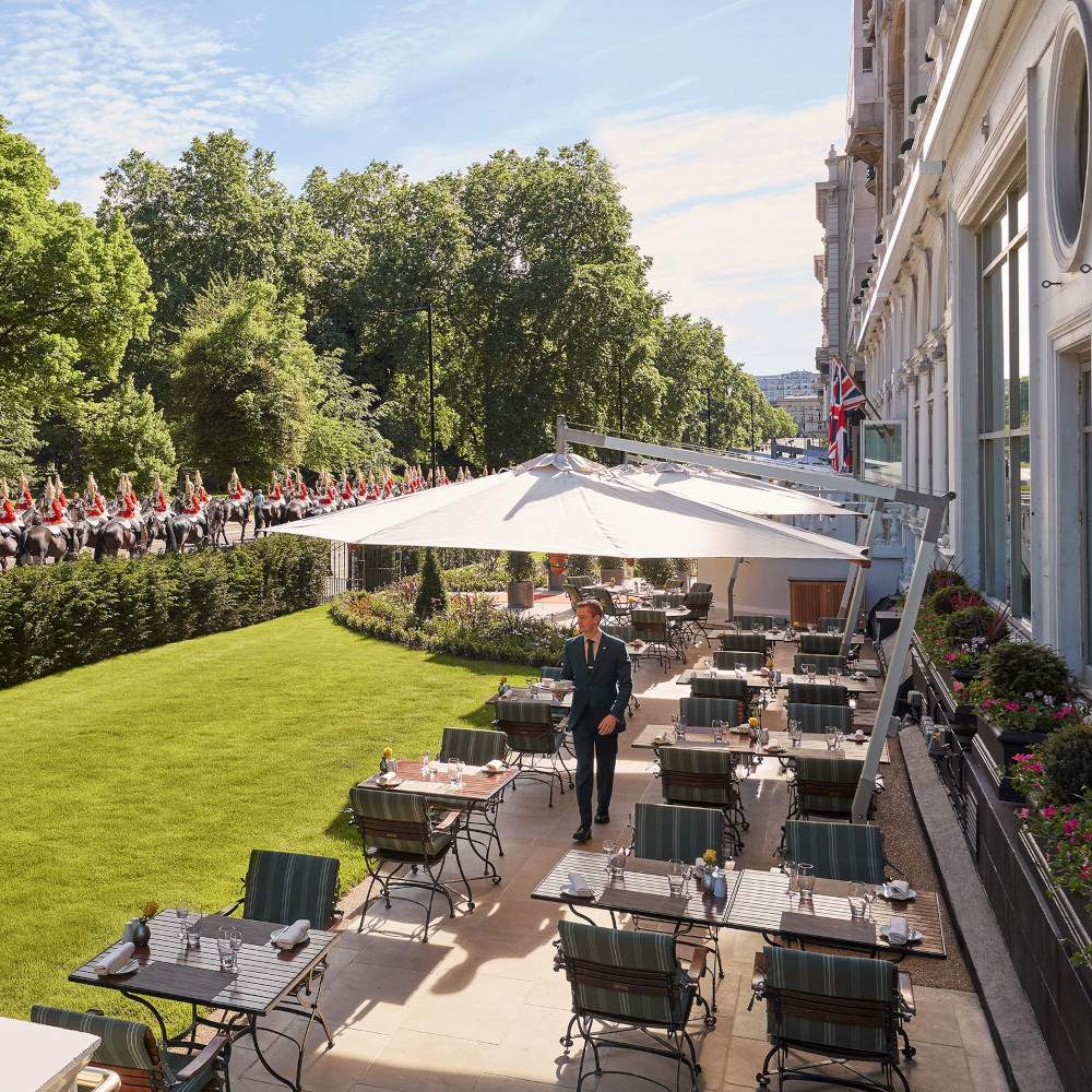 An outdoor patio seating area with tables and umbrellas overlooking a small lawn and, beyond, British guards on horses.