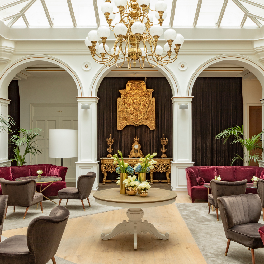 Bright interior of a lobby with roof windows, a large chandelier, arched doorways and red plush sofas.