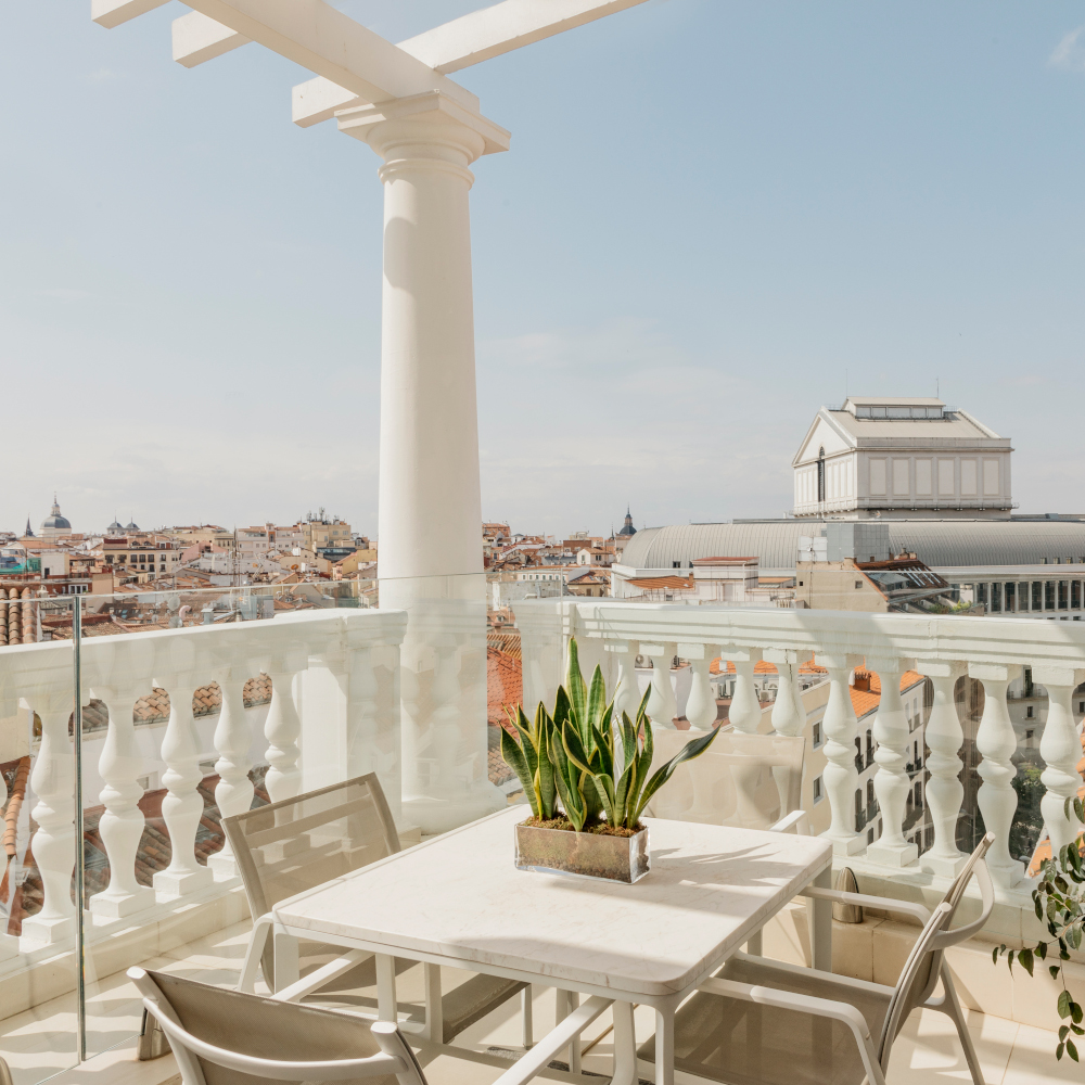 Exterior of a balcony with white pillar and railing and two chairs overlooking the rooftops and buildings of Madrid.