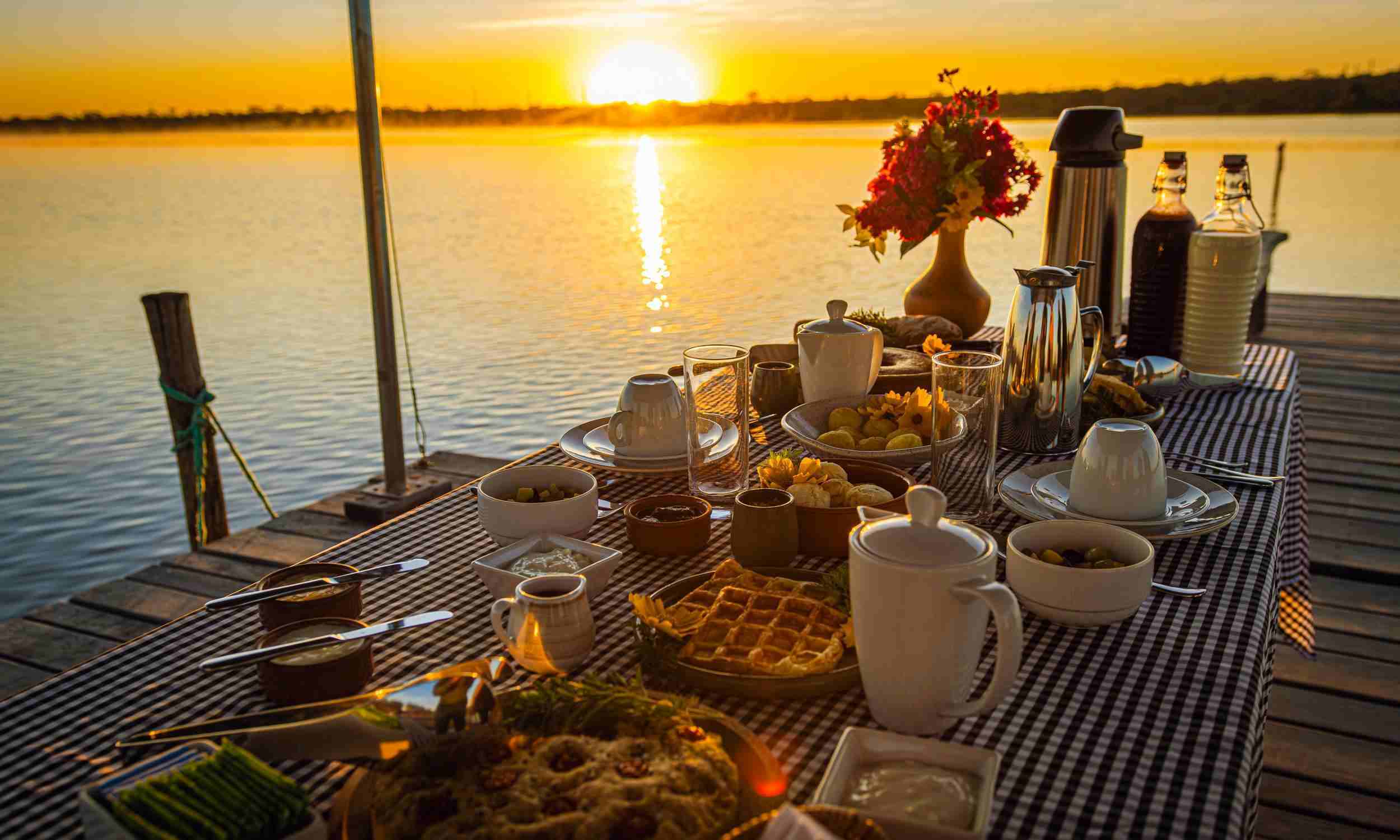 A table laid with a blue and white checked tablecloth and set with breakfast food and coffee, on a pier jutting over a lake as the sun rises in the background.