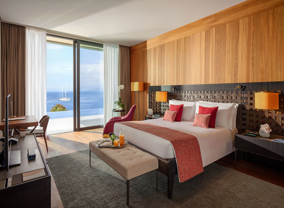 A double bed with white sheets and red cushions and throw, with a wooden headboard and footstool laid with a breakfast tray. Sliding glass doors frame views of the sea and a yacht in the distance.