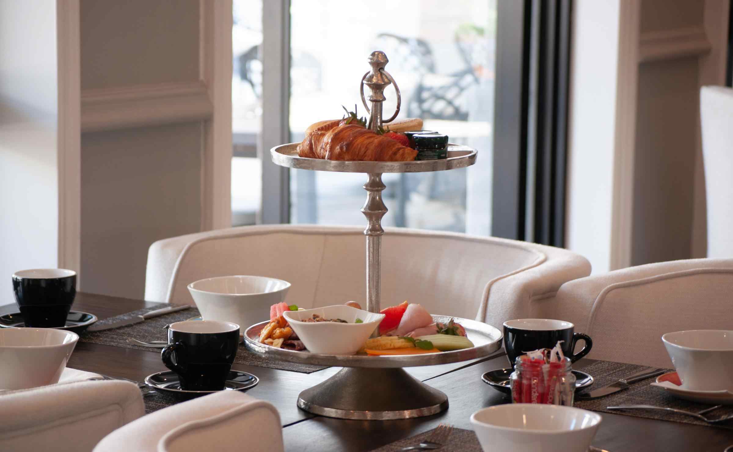 A close-up view of a table set for breakfast, with cream armchairs, cups and saucers and a cake stand with croissants, fruit and jams. Glass doors in the background lead to a terrace.